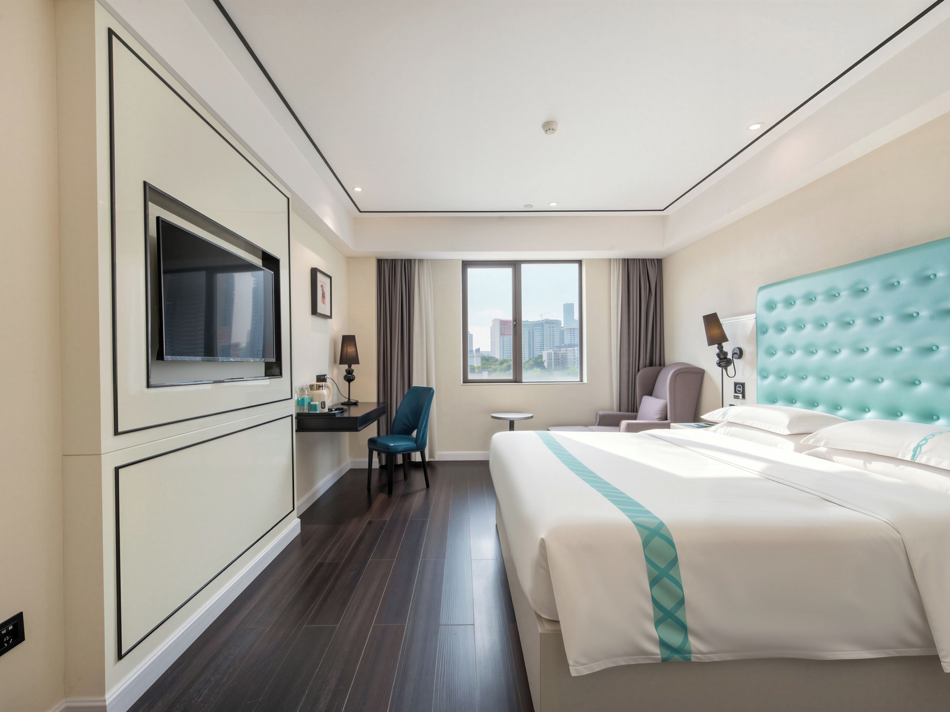 A hotel room with a large bed, a desk with a chair, a television, and a view of the city.