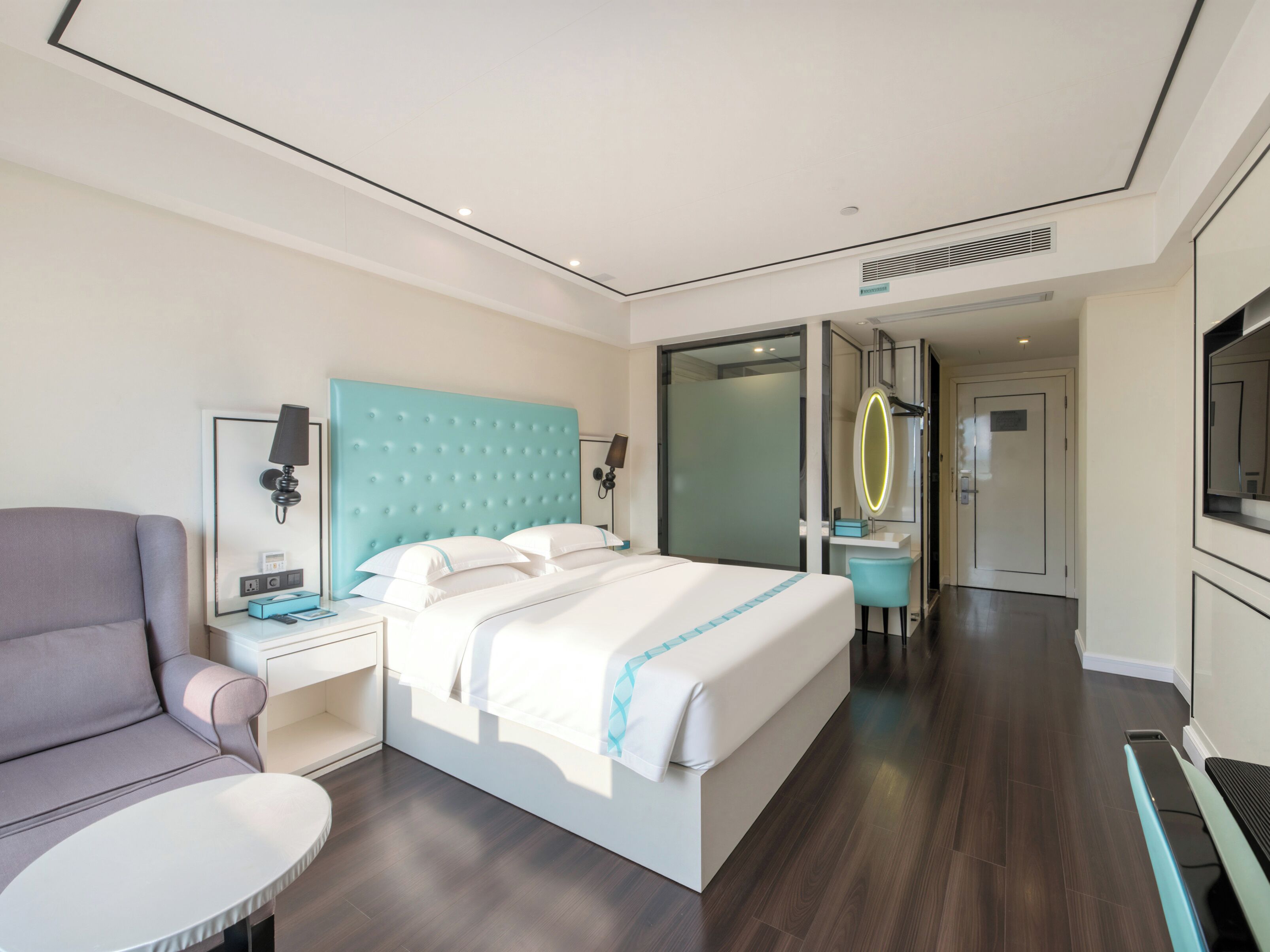 Una habitación de hotel moderna con una cama grande, un área de descanso con una silla, un escritorio con lámpara y un baño con espejo y lavamanos.