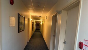 A hotel hallway with a patterned carpet, framed artwork on the walls, and a fire alarm on the right. - Fairview Inn (Simcoe)
