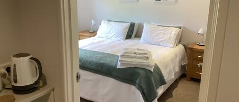 A neatly made bed with white and green bedding, a stack of folded towels, and a white kettle on a bedside table.