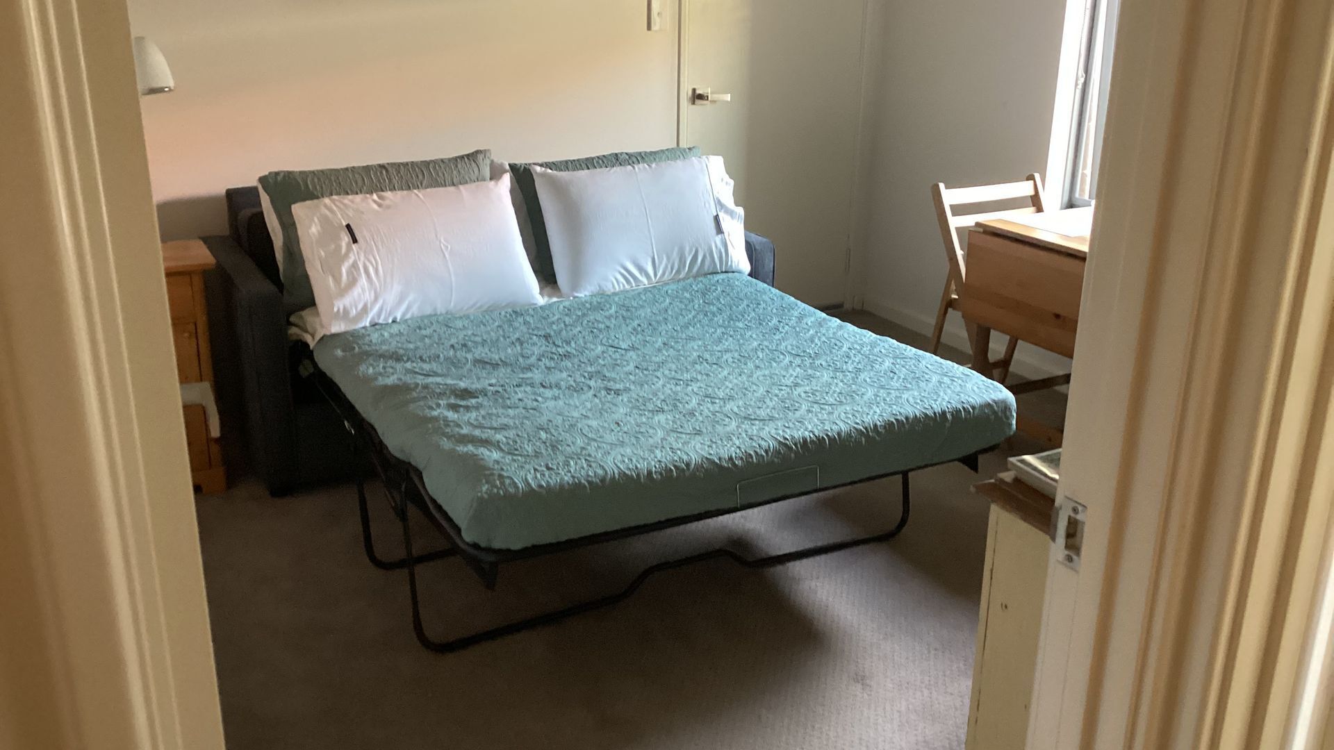 Una cama individual con colcha azul y dos almohadas, una mesita de noche de madera, un escritorio con silla, y una puerta.