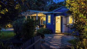 A house with a covered porch, a stone pathway leading to the entrance, and a well-maintained garden. - Totara Cottage - Our Island (Waiheke Island)