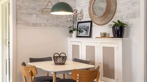 A dining area with a white table, four wooden chairs, a woven basket, a green pendant light, a wicker mirror, and a potted plant. - Totara Cottage - Our Island (Waiheke Island)