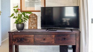 A wooden TV stand with a flat-screen TV, a potted plant, a remote control, and a decorative sign. - Totara Cottage - Our Island (Waiheke Island)