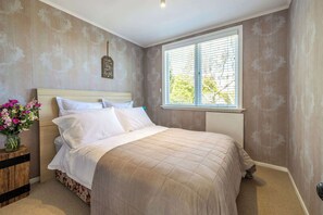 A bedroom with a bed, a nightstand with a vase of flowers, and a window with blinds. - Totara Cottage - Our Island (Waiheke Island)