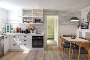 A modern kitchen with white cabinetry, a built-in oven, and a dining area with a table and chairs. - Totara Cottage - Our Island (Waiheke Island)