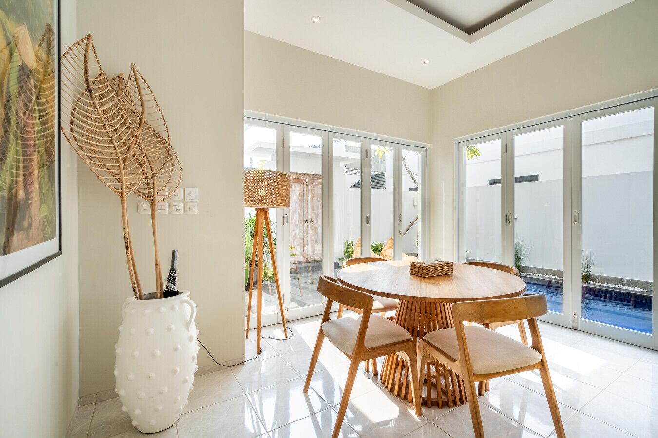 A modern dining area with a round table and wooden chairs, large windows, and a view of a swimming pool.