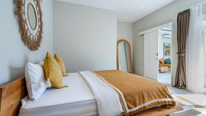 A bedroom with a wooden bed, white and yellow pillows, a brown throw blanket, a wall mirror, and a view into another room through an open door.
