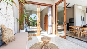 A modern room with a sofa, a dining table, and a view through a glass door to an outdoor area with plants. - Villa Glentana - 3BR EPIC Sunset Villa (Seseh)