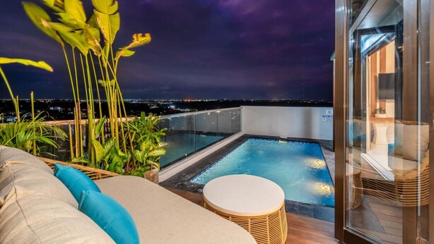 A rooftop pool area with a glass balustrade, lounge seating, and a view of the city at night.