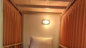 Una cama individual con un edredón blanco y una toalla blanca enrollada, una lámpara redonda de pared y paneles de madera en las paredes.