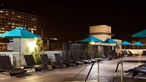 Un espace piscine sur le toit, avec des chaises longues et des parasols, un jacuzzi et une vue sur le paysage urbain illuminé la nuit.
