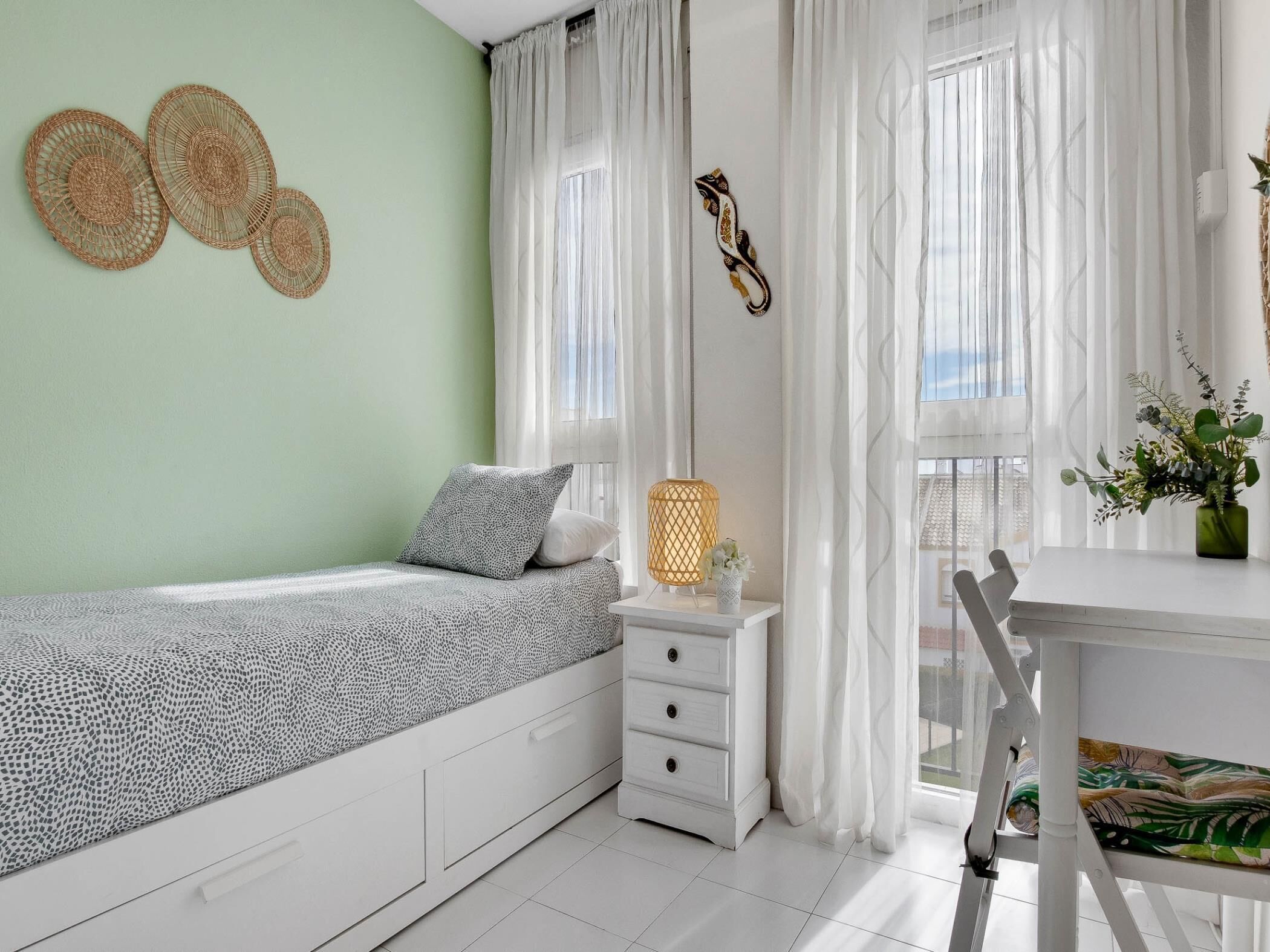 A bedroom with a bed, a bedside table with a lamp, a chair, and a window with curtains.
