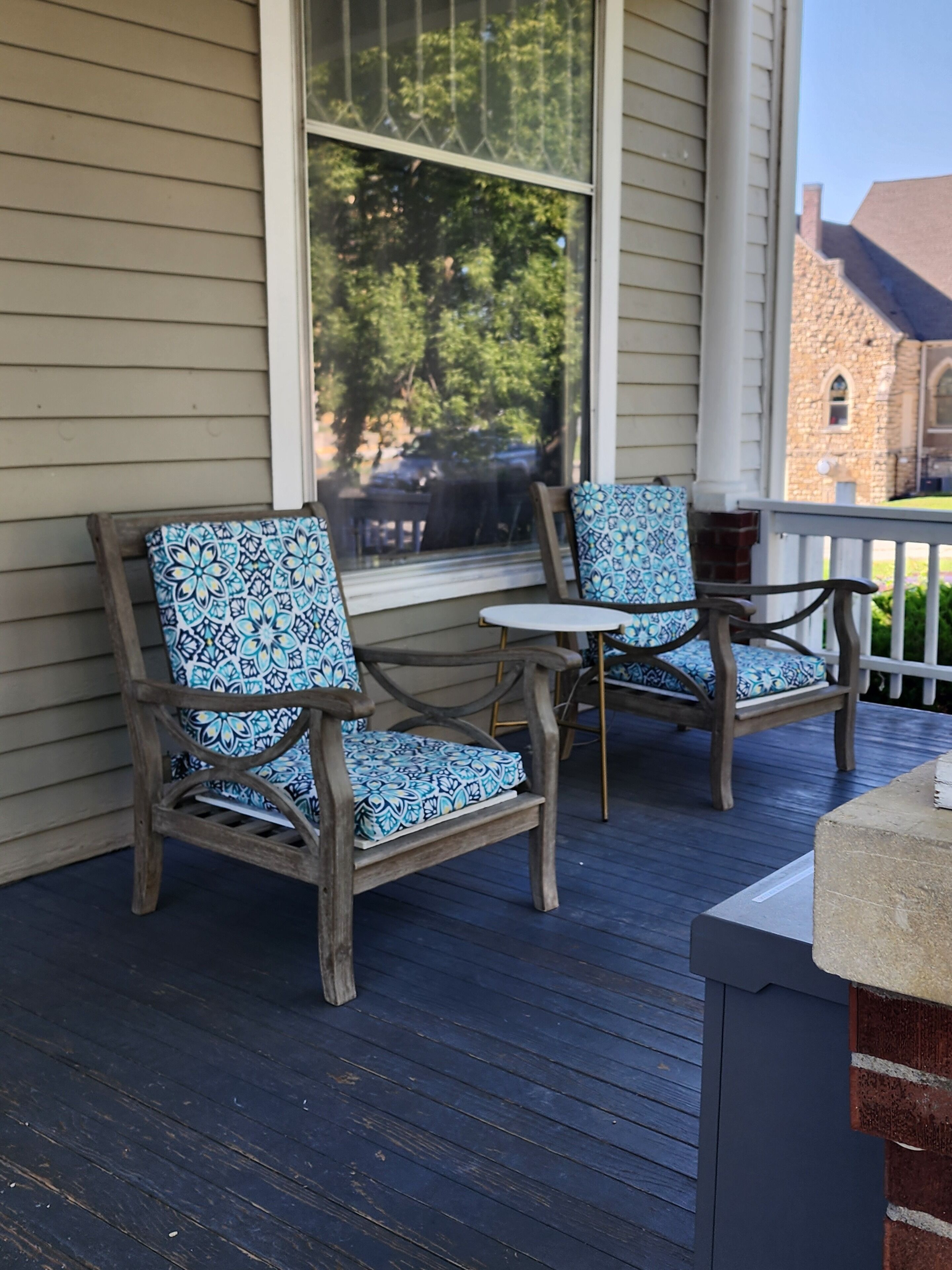A wooden porch with two chairs and a small table, a window, and a stone building in the background.