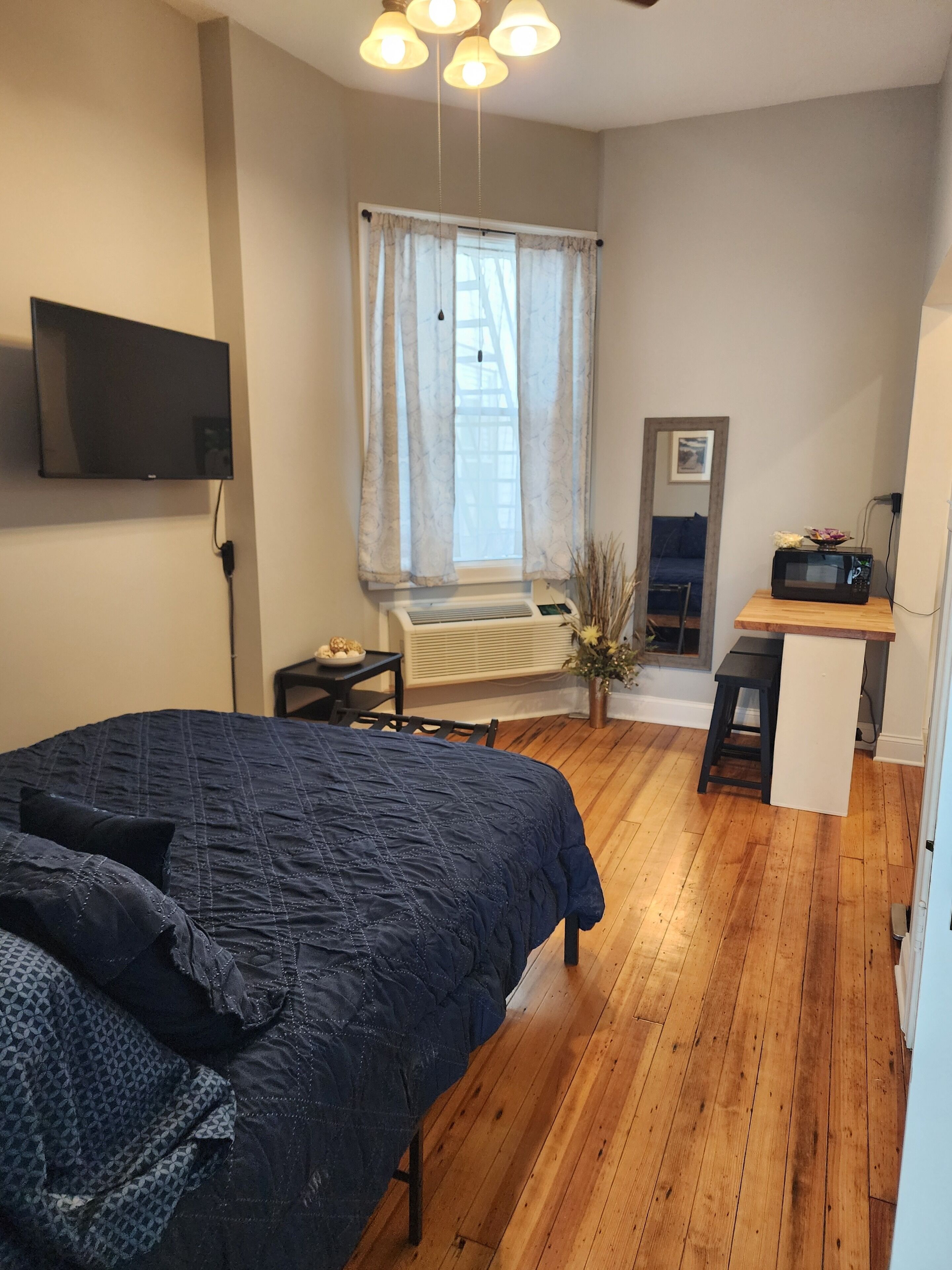Une chambre à coucher avec un lit, un téléviseur fixé au mur, une petite table, un four à micro-ondes et une fenêtre avec des rideaux.