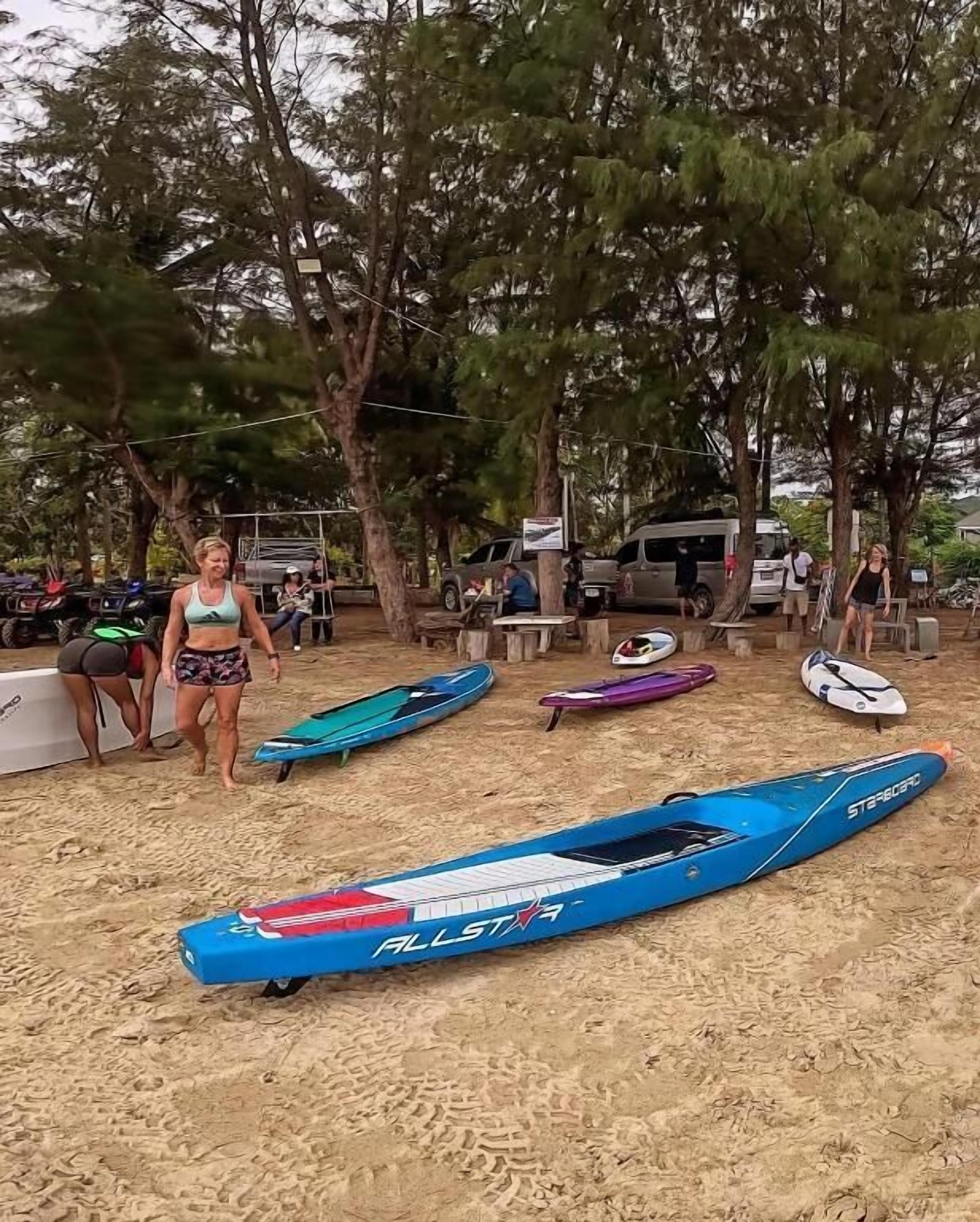 Une plage de sable avec plusieurs planches de surf, quelques personnes et des véhicules en arrière-plan.