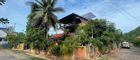 A two-story house with a red roof, surrounded by greenery and a palm tree.