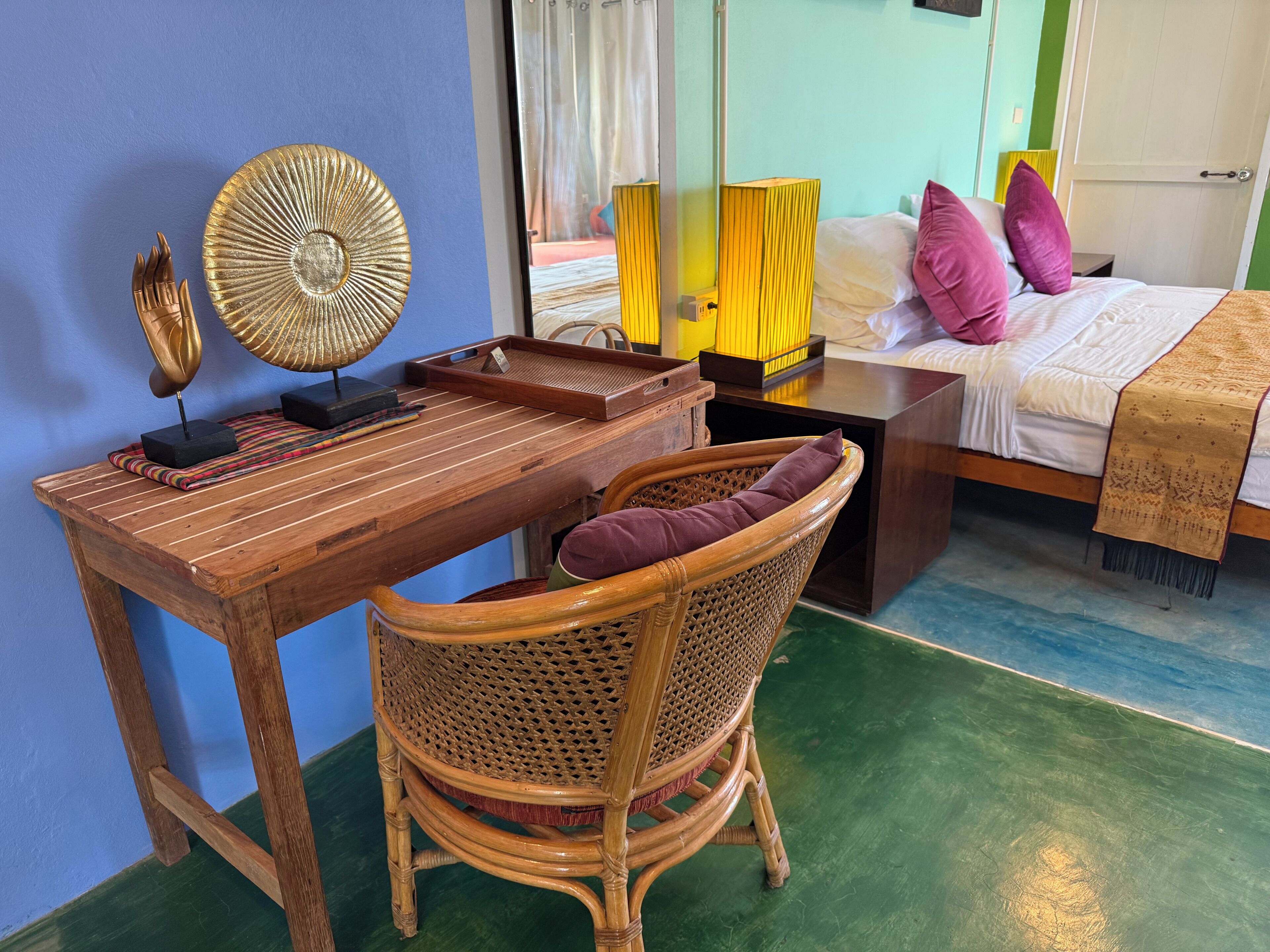 A room with a wooden desk, a wicker chair, a bed with pillows, and a mirror reflecting a room with curtains.