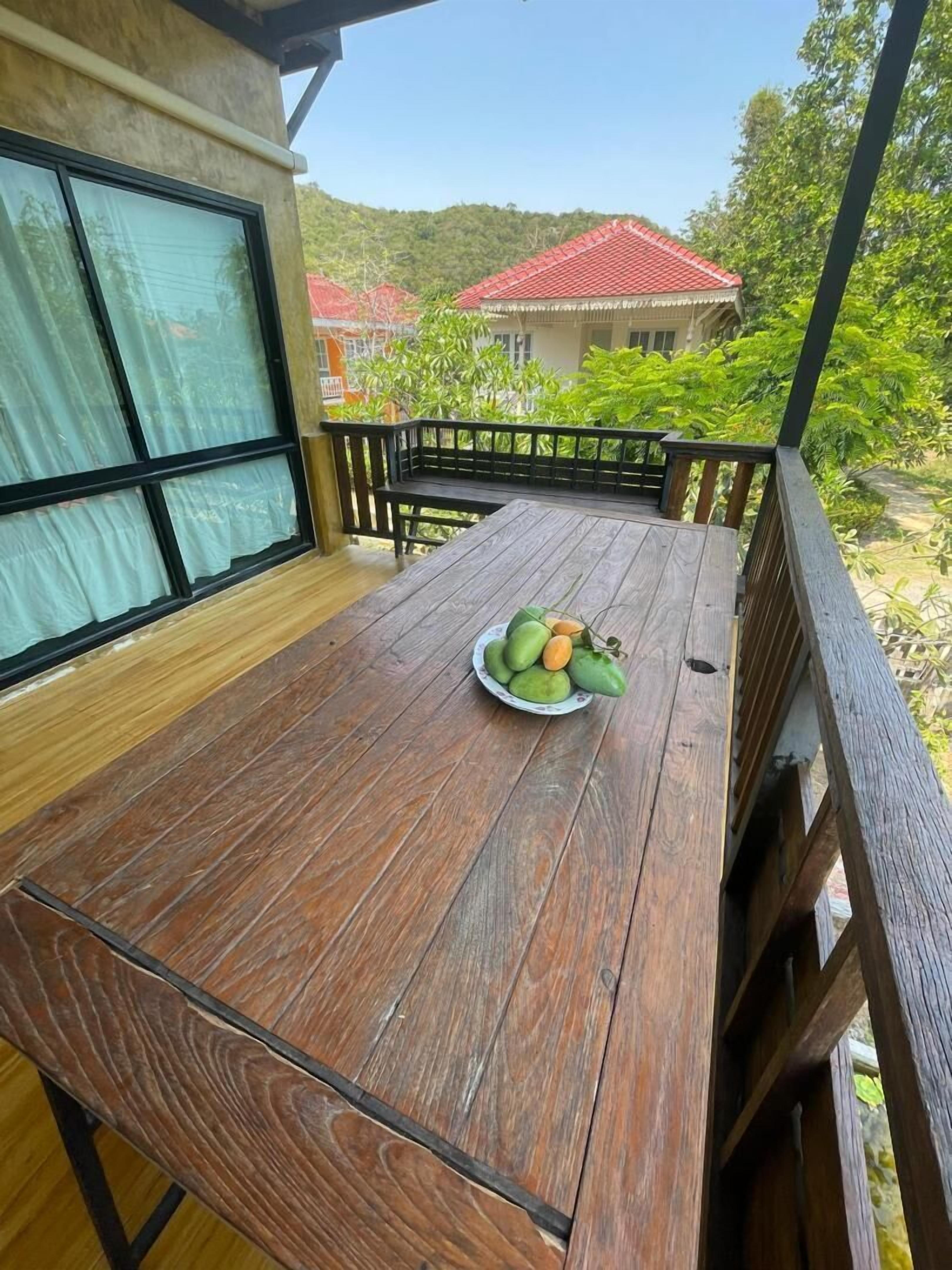 Une terrasse en bois avec une table, un saladier rempli de fruits et une vue sur des maisons et de la verdure.