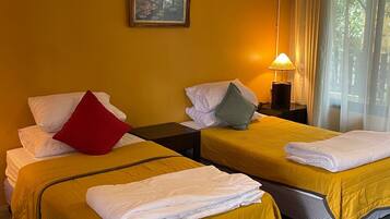 Een hotelkamer met twee bedden, elk met een gele sprei, witte kussens, en een rood kussen op één bed. Tussen de bedden staat een nachtkastje met een lamp, en aan de muur hangt een lijst met een afbeelding.