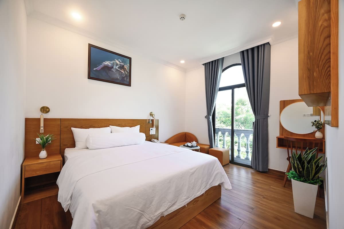 Una camera d'albergo con un letto grande, una testiera in legno, un comodino con una lampada, un tavolino con una pianta e un'ampia finestra con tende.