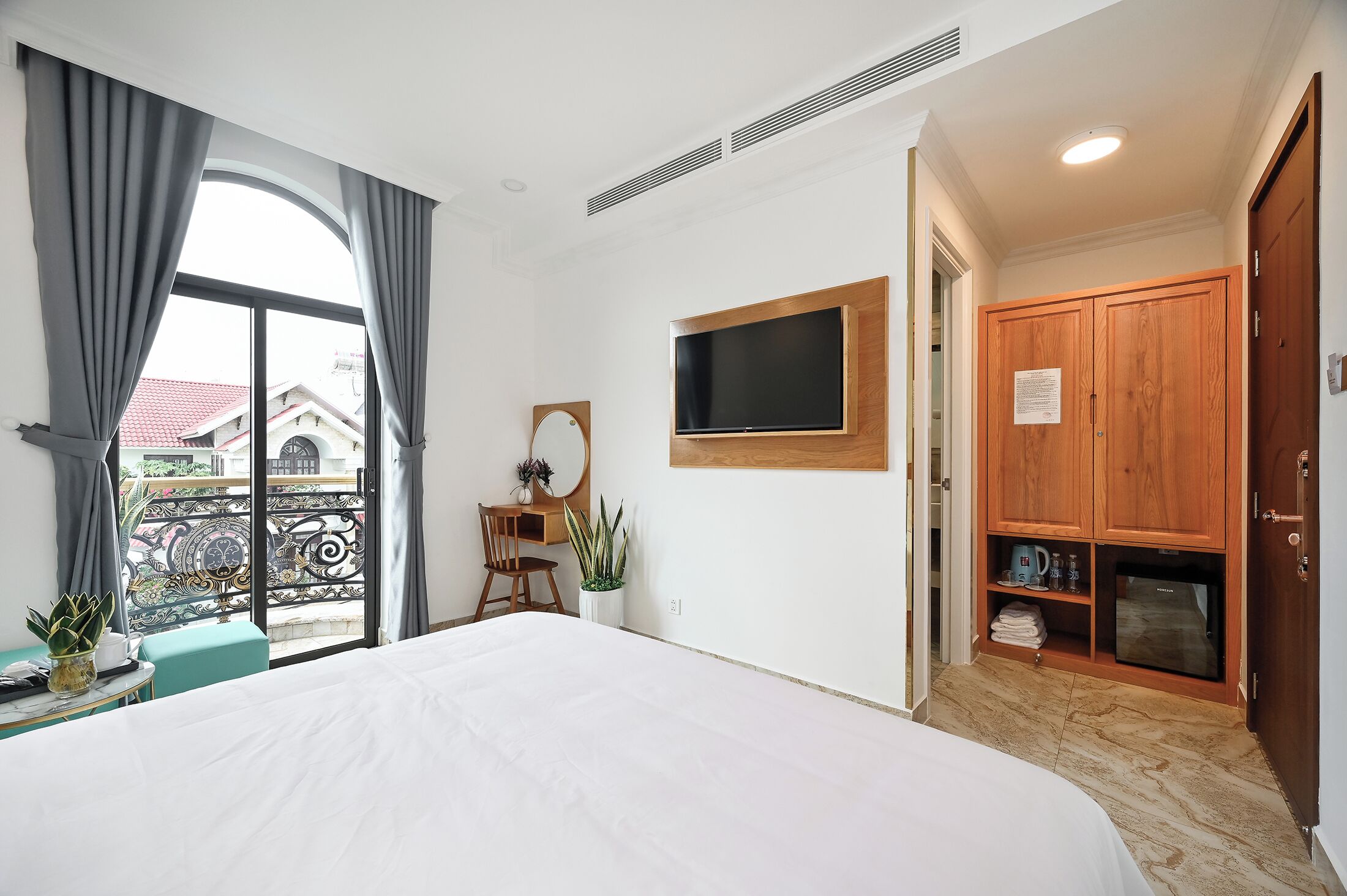 Habitación de hotel con cama, escritorio con silla, televisor montado en la pared, armario y balcón con vistas a edificios.