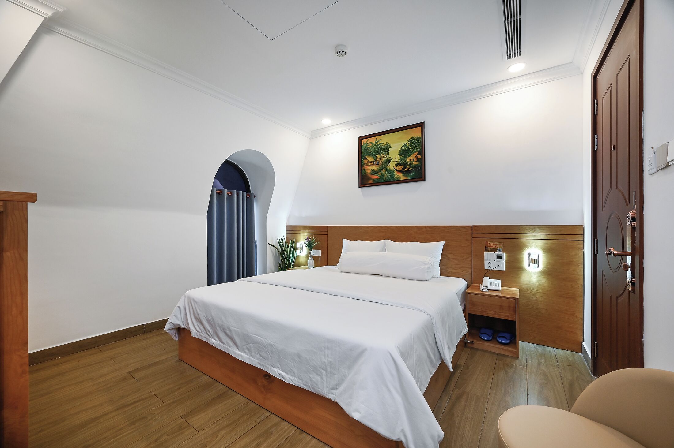A hotel room with a large bed, a wooden headboard, a nightstand with a lamp, a small table with a phone, and a door with a handle.
