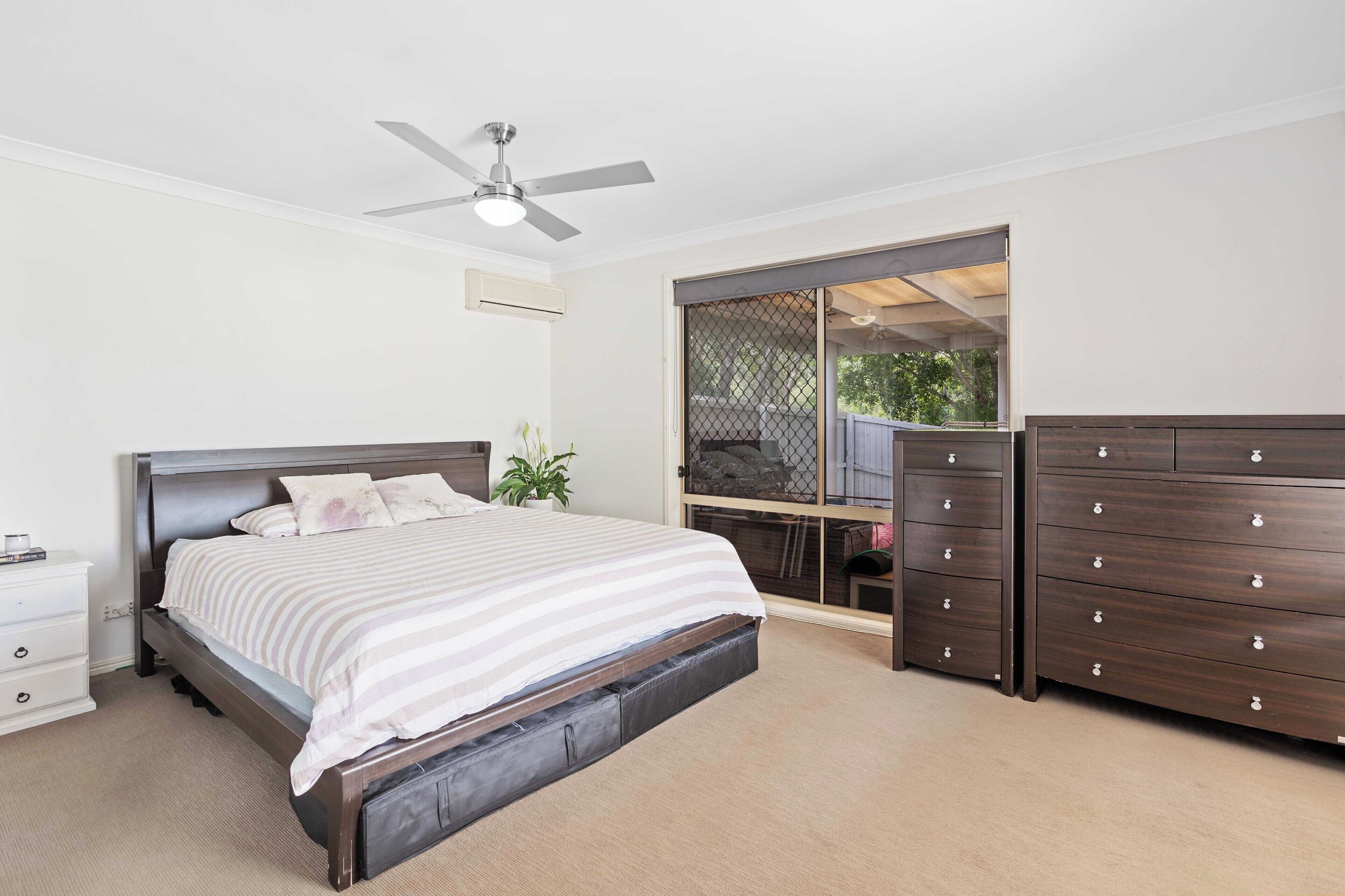 A bedroom with a bed, a dresser, a ceiling fan, and a window with a view of the outdoors.