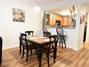 A dining area with a wooden table and chairs, a kitchen in the background, and a painting on the wall.