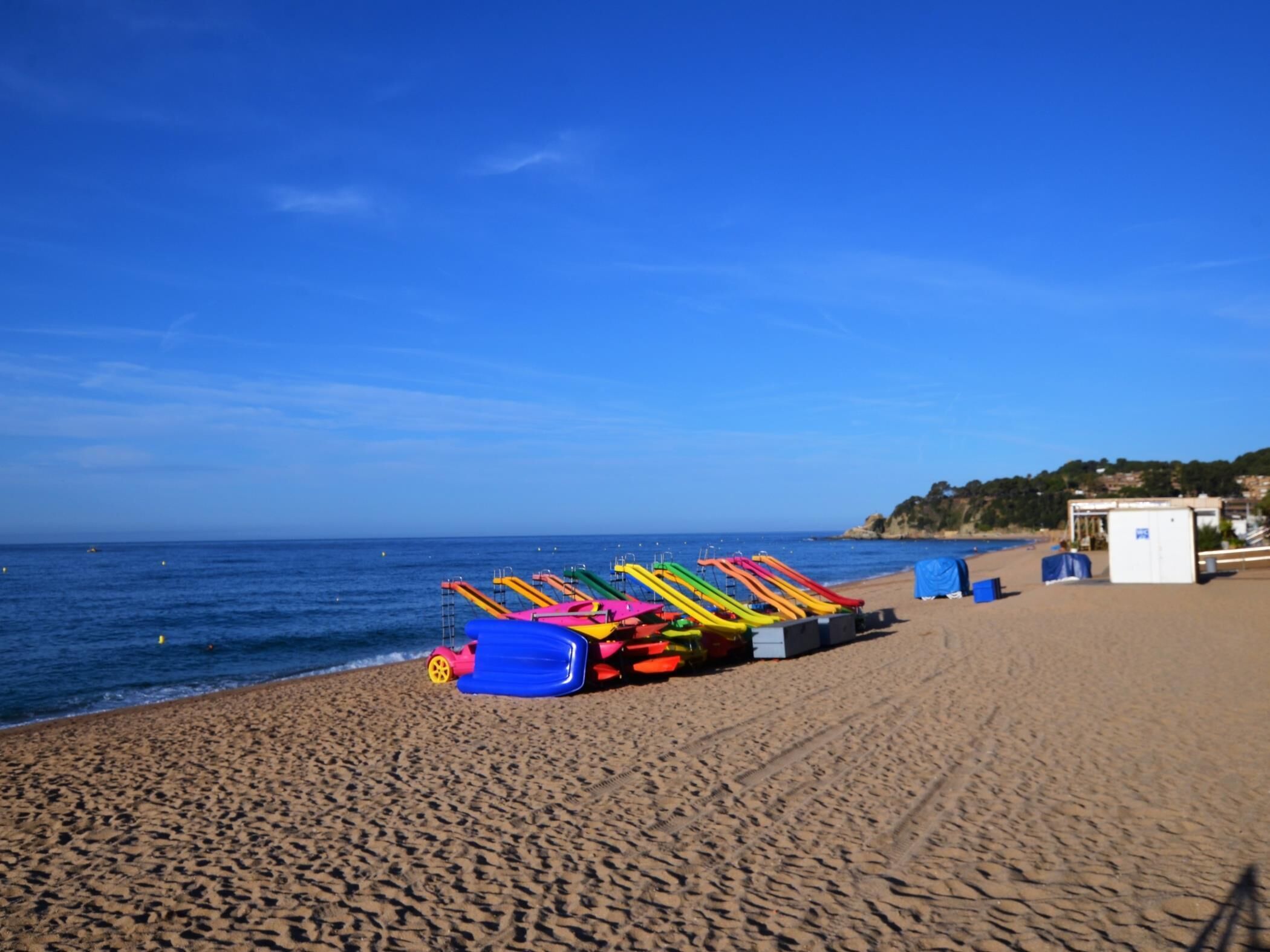 Une plage bordée d’une rangée de kayaks aux couleurs vives, avec un petit bâtiment en arrière-plan, qui semble être un poste de surveillance des sauveteurs.