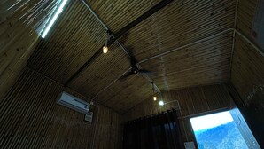 Een kamer met een bamboe plafond, een bed met wit beddengoed, een airconditioning en uitzicht op een bergachtig landschap door een raam.