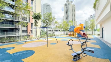 An outdoor playground with swings, slides, and climbing structures, surrounded by high-rise residential buildings.