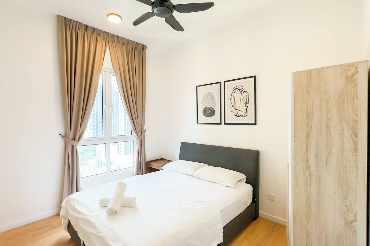 A bedroom with a bed, a ceiling fan, a window with curtains, and two framed artworks on the wall.