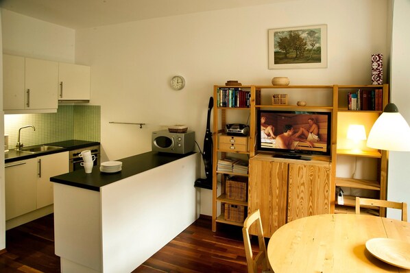 A compact kitchen with a dining area, a wooden bookshelf, and a television.