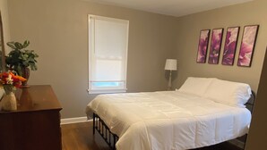 A bedroom with a wooden floor, a bed with white bedding, a dresser, a window with blinds, and four wall-mounted flower paintings.