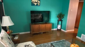 A living room with teal walls, a sofa with floral upholstery, a glass coffee table, a wooden entertainment center, and a flat-screen TV.
