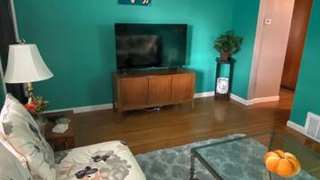 Un salon aux murs de couleur turquoise, un canapé recouvert d’un tissu à motifs floraux, une table basse en verre, un meuble de rangement en bois pour appareils électroniques et un téléviseur à écran plat.