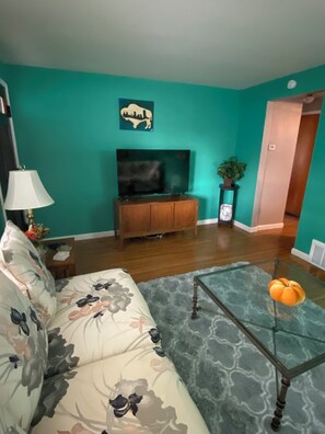 A living room with teal walls, a sofa with floral upholstery, a glass coffee table, a wooden entertainment center, and a flat-screen TV.