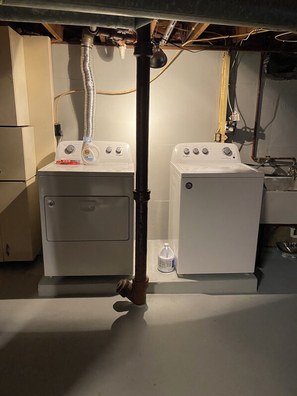 Two front-loading washing machines under a house's basement ceiling, with visible plumbing and electrical wiring.