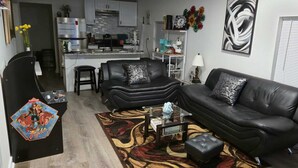 A modern living room with a black leather sofa, a glass coffee table, and a patterned rug. There is a kitchen in the background with white cabinets and a stainless steel refrigerator.