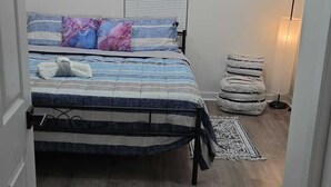 A bedroom with a bed, a ceiling light fixture, a window with blinds, a wall-mounted shelf with decorative items, and a floor lamp.