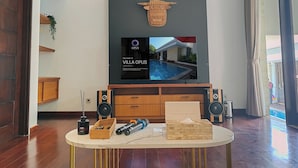 Un salón moderno con un televisor grande mostrando una villa, paneles de madera, una mesa redonda con un cuaderno y un altavoz.
