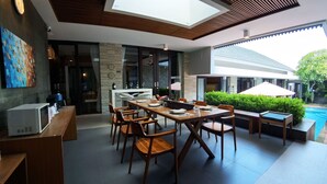 Un comedor moderno con una mesa de madera larga y sillas, una zona de cocina con encimera blanca, y vistas a una piscina y vegetación exterior.