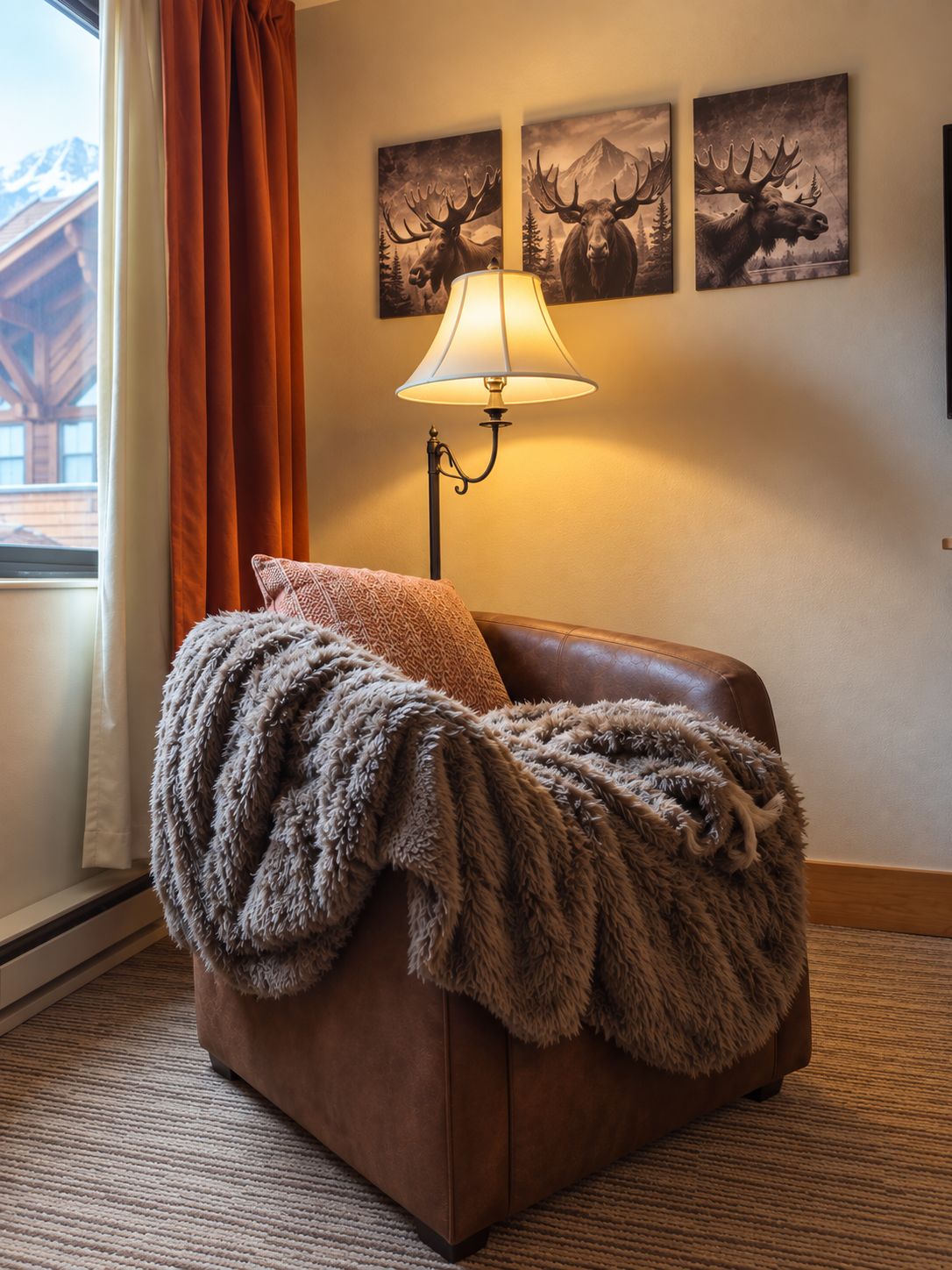 A cozy room with a brown leather sofa, a fluffy throw blanket, and a lamp. There are three moose-themed paintings on the wall.