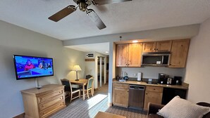Un espacio habitable compacto con comedor, kitchenette y un televisor mostrando un noticiero.