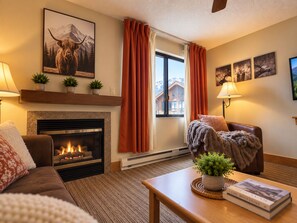 Un salon chaleureux avec une cheminée, un canapé, une table basse avec un livre et une fenêtre avec des rideaux.