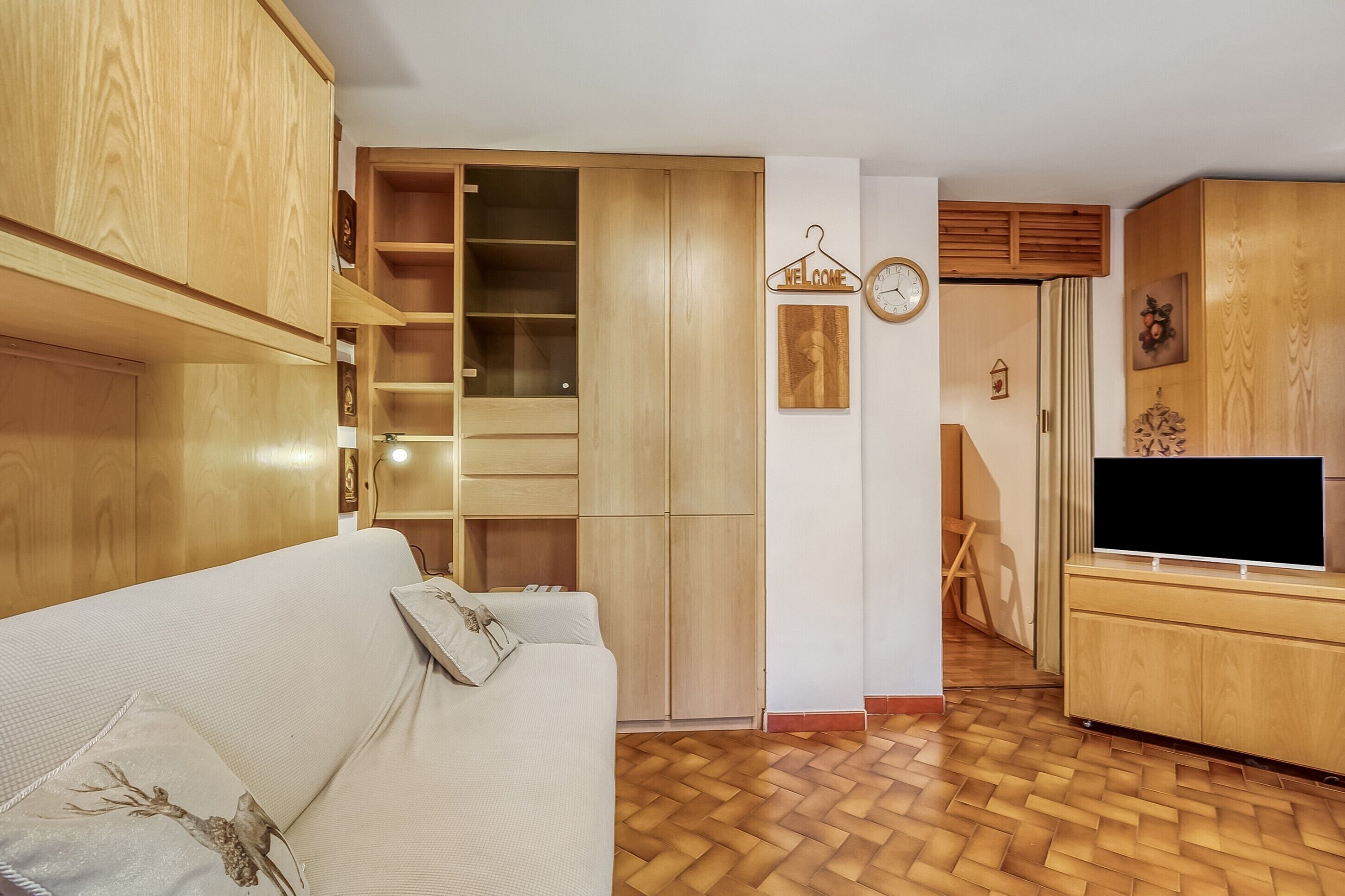 Una habitación con un armario de madera, un sofá blanco, un televisor sobre un mueble de madera y suelo de madera.