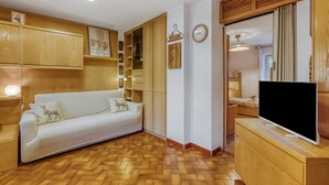 Een kamer met een witte bank, houten kasten, een televisie op een houten standaard, een klok en een hangend bord met de tekst 'Welcome'.