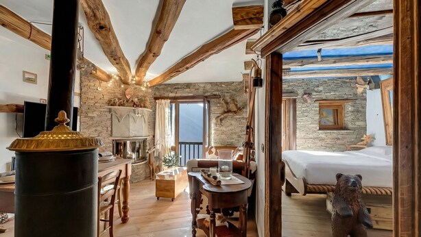 A cozy room with a stone wall, wooden beams, a bed with a bear decoration, a dining table, and a fireplace. - Grange Romantic Retreat Mountain View (Sauze di Cesana)