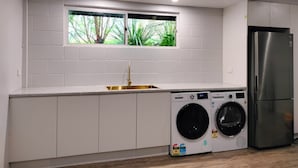 A compact kitchen with a built-in sink, a window with a view of greenery, a stainless steel refrigerator, and a front-loading washing machine.